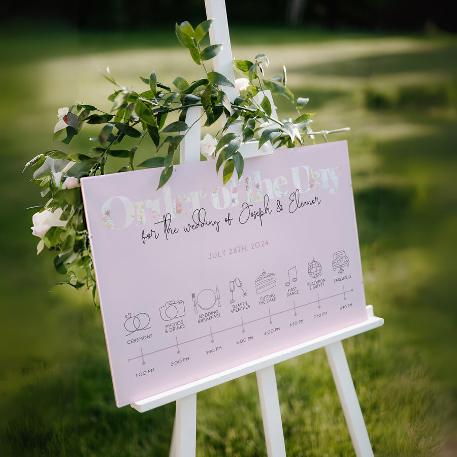 pastel pink wedding order of the day sign, with beautiful calligraphy text and a floral white header.